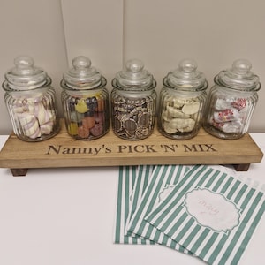 May include: Five glass jars with lids filled with sweets, displayed on a wooden tray that reads "Nanny's PICK 'N' MIX". The jars contain marshmallows, jelly sweets, chocolate coins, banana sweets, and heart-shaped sweets. Green and white striped paper bags are in the foreground.
