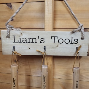 May include: A wooden sign with the text "Liam's Tools" in black lettering. The sign has three metal hooks with leather straps, and a rope for hanging. The background is a light brown wooden wall.