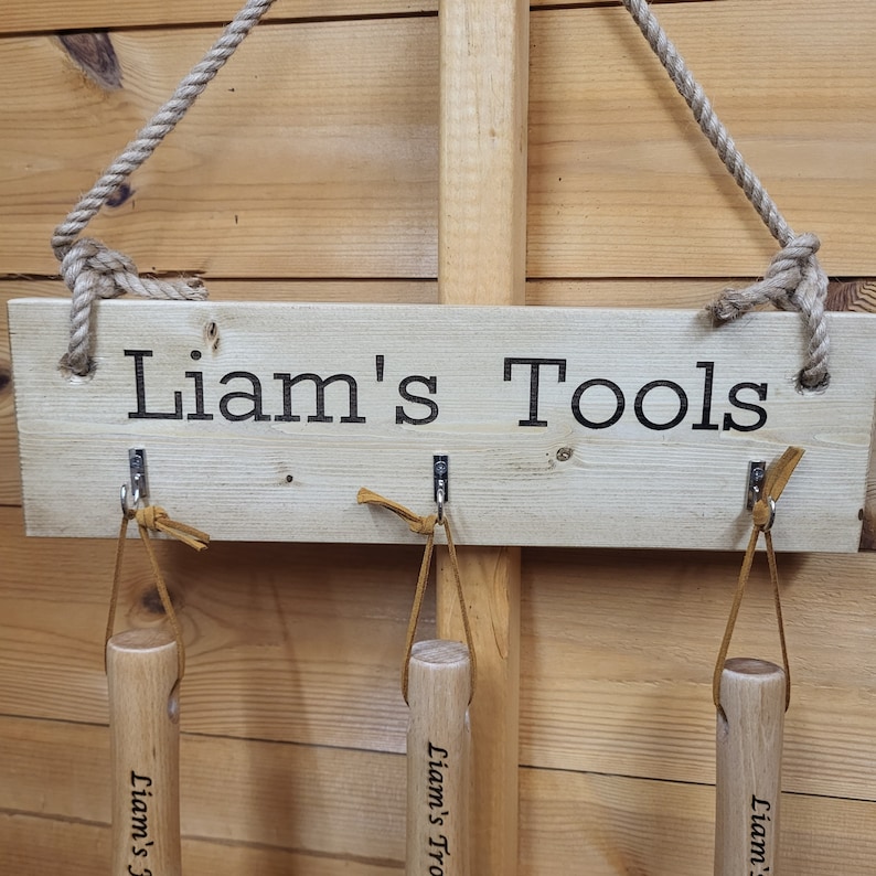May include: A wooden sign with the text "Liam's Tools" in black lettering. The sign has three metal hooks with leather straps, and a rope for hanging. The background is a light brown wooden wall.