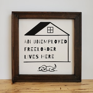 May include: Square wooden framed sign with the text "AN UNEMPLOYED FREELOADER LIVES HERE" in black letters. A black illustration of a house and a dog are also on the sign. The frame is dark brown.
