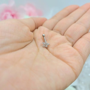 May include: A silver belly button ring with a flower design. The flower is made of small, clear gemstones. The top ball is also silver. The jewelry is small and delicate.