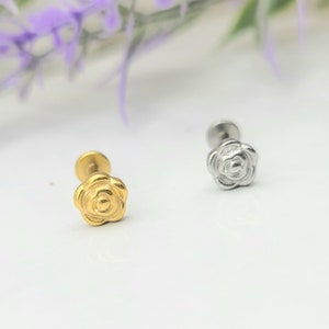 May include: Two small, flower-shaped earrings. One is gold-colored, the other is silver-colored. The earrings have a flat back and a post for piercing. The earrings are on a white surface with purple flowers in the background.