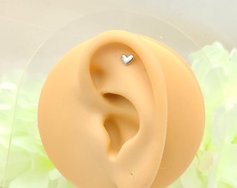 May include: A small, silver heart-shaped earring stud is displayed on a tan ear model. The earring is positioned on the upper ear cartilage. The background is a soft, blurred mix of white and green.
