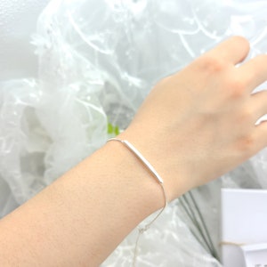 May include: A silver bracelet featuring a slender, rectangular bar design. The bracelet is delicate and appears to be made of a shiny metal. The bar is centered on a thin chain, creating a minimalist and modern aesthetic. The bracelet is shown on a wrist.