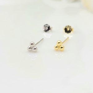 May include: A pair of stud earrings, each with three small, round beads. One earring is silver, the other is gold. Two earring backs are also visible. The earrings are on a white surface.