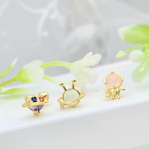 May include: Three gold-coloured stud earrings with space-themed designs. One features a planet with colourful gems, another a planet with a light green orb, and the last an astronaut with a pink gem head. Each earring has small, clear gems.