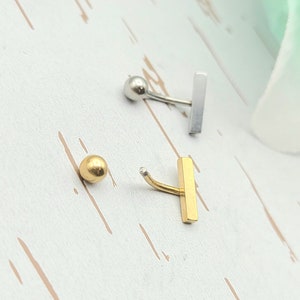 以下が含まれることがあります： 2つのカーブしたバーベルボディジュエリー。1つはボールエンドと長方形のバーが付いたシルバーカラー。もう1つはボールエンドと長方形のバーが付いたゴールドカラー。ジュエリーは白い表面にあります。