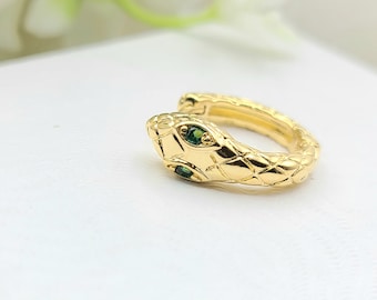 Puede incluir: Un pendiente de oreja dorado con forma de serpiente. La cabeza de la serpiente tiene dos gemas verdes como ojos. El pendiente tiene un patrón texturizado en forma de escamas. La joya está sobre un fondo blanco suave.