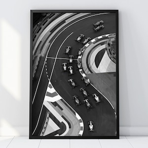 Puede incluir: Una impresión de arte mural enmarcada en blanco y negro que presenta una vista aérea de varios coches de carreras navegando por una sección de pista curva. La composición captura los vehículos en movimiento sobre el asfalto.