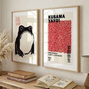 Può includere: Due stampe artistiche incorniciate in legno chiaro. La stampa a sinistra mostra un'illustrazione di una rana in stile inchiostro nero su sfondo crema con un sigillo rosso. Quella a destra presenta pois rossi, il testo KUSAMA YAYOI e caratteri giapponesi.