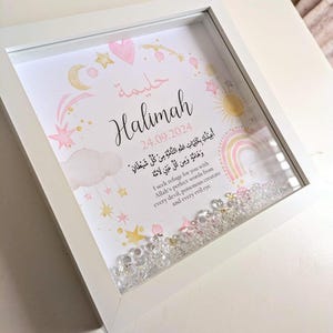 May include: A white frame with a pink and gold design featuring a name, date, and Arabic text. The frame is filled with clear glitter.