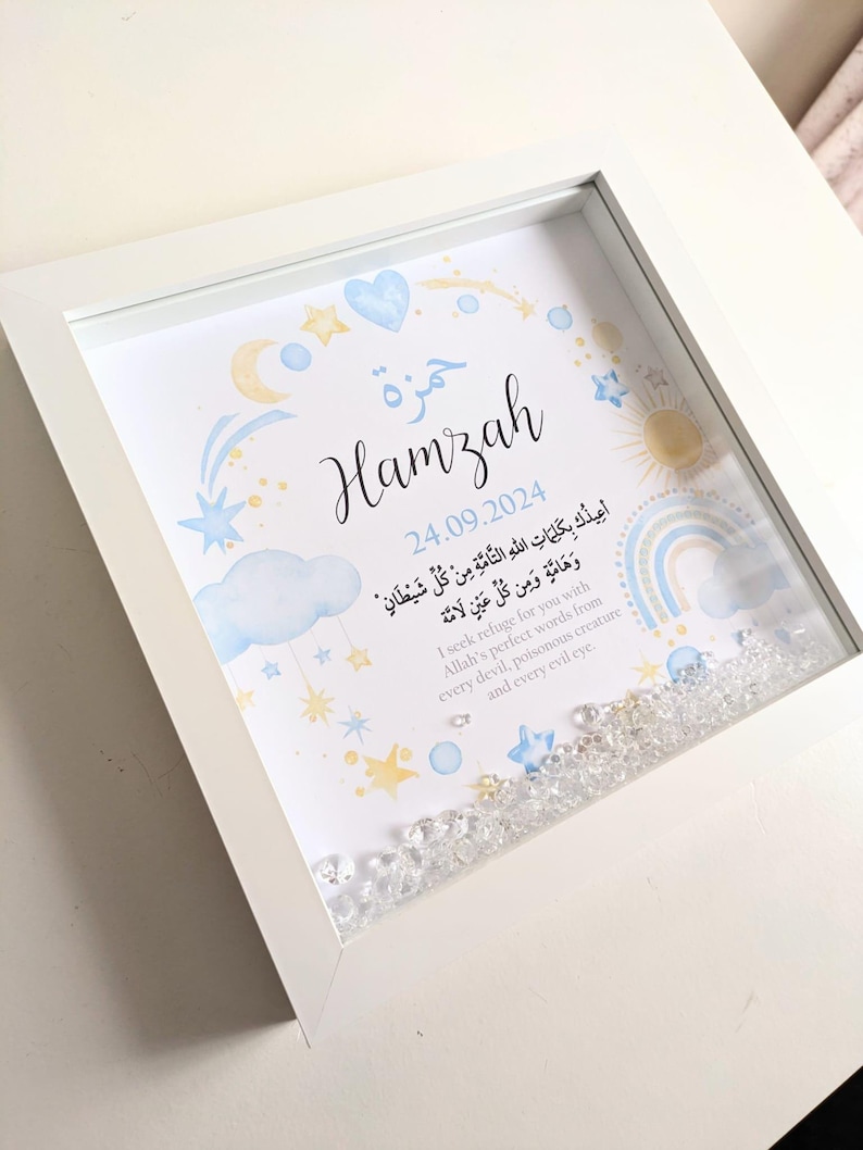 May include: A white frame with a blue and yellow watercolor illustration of stars, a crescent moon, a rainbow, and a sun. The name "Hamzah" is written in Arabic calligraphy and the date "24.09.2024" is written below. The frame is filled with clear crystals. The text "أعيدكَ بِكَلِمَاتِ اللَّهِ التَّامَّةِ مِنْ كُلِّ شَيْطَانٍ وَهَامَّةٍ وَمِن كُلِّ عَيْنٍ لَامَّة" is written in Arabic script. The text "I seek refuge for you with Allah's perfect words from every devil, poisonous creature and every evil eye." is written in English below the Arabic text.