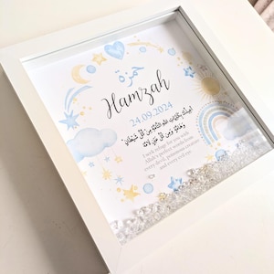 May include: A white framed picture with a blue and yellow watercolor design. The name Hamzah is written in Arabic calligraphy. The date 24.09.2024 is written below the name. The text in Arabic translates to "I seek refuge for you with Allah's perfect words from every devil, poisonous creature and every evil eye."