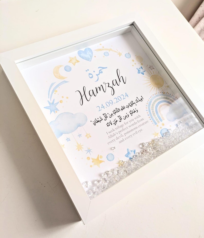 May include: A white framed picture with a blue and yellow watercolor design. The name Hamzah is written in Arabic calligraphy. The date 24.09.2024 is written below the name. The text in Arabic translates to "I seek refuge for you with Allah's perfect words from every devil, poisonous creature and every evil eye."