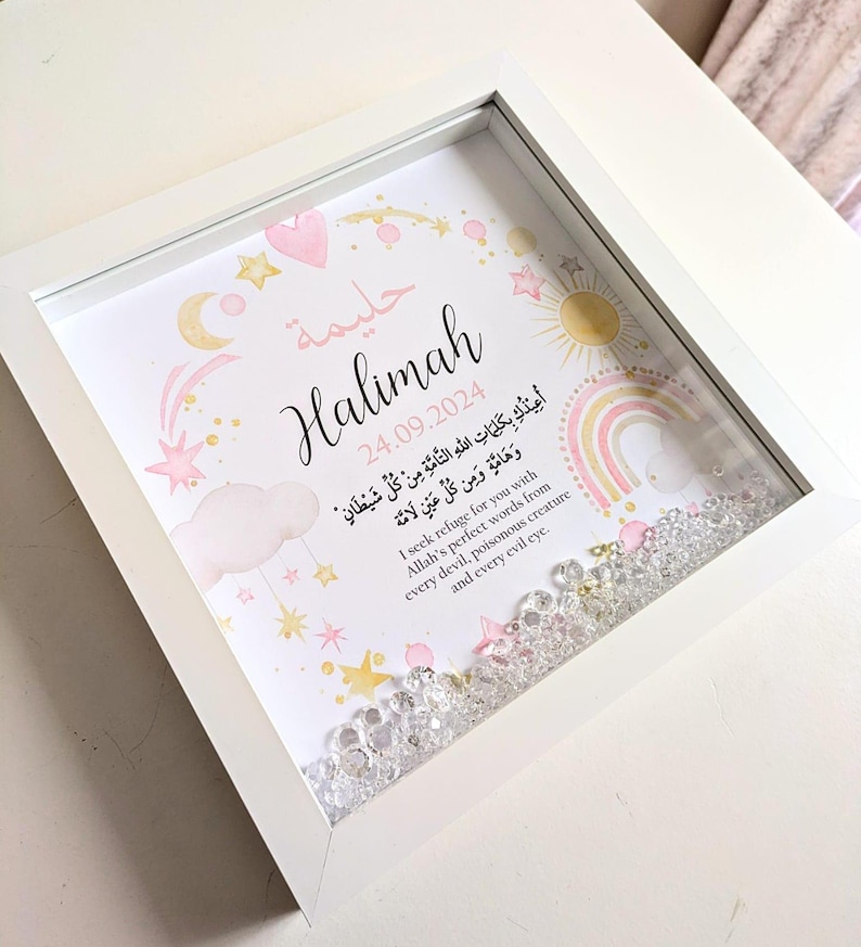May include: A white frame with a pink and gold design. The frame contains a printed image with the name "Halimah" and the date "24.09.2024". The image also includes a rainbow, stars, and clouds. The frame is filled with clear crystals.