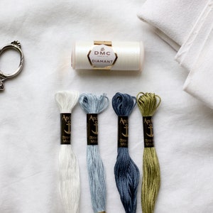 Könnte beinhalten: Ein Set mit Stickzubehör, darunter weißer Aida-Stoff, eine Spule weißer DMC Diamant Stickgarn, eine silberne Stickerschere und vier Spulen Anchor Stickgarn in weiß, hellblau, dunkelblau und grün.