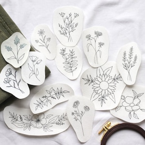 Könnte beinhalten: Ein Set mit 12 Stickmustern mit verschiedenen floralen Designs, darunter Sonnenblumen, Lavendel und andere Wildblumen. Die Muster sind auf weißem Papier gedruckt und bereit zum Sticken.