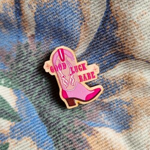 Puede incluir: Un pin de madera con forma de bota de vaquero rosa con la inscripción "GOOD LUCK BABE". La bota tiene un tacón marrón y costuras decorativas. El pin está sobre un fondo estampado.