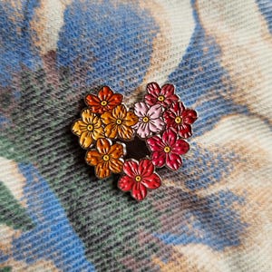 Peut inclure: Une épingle en émail en forme de cœur représentant un groupe de fleurs colorées. Les fleurs passent de l'orange au rose et au rouge, avec des centres jaunes. L'épingle a une bordure en métal argenté.