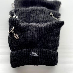 May include: Dark grey knitted beanie hats with silver metal details. Each hat has a small charm, including a razor blade. The label on the hat reads "OTHERS MADE IN HELL".