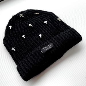 Pode incluir: Gorro de malha preto com várias cruzes prateadas. O gorro apresenta um design canelado e uma pequena etiqueta que diz "OTHERS MADE IN HELL". Um acessório elegante e arrojado.