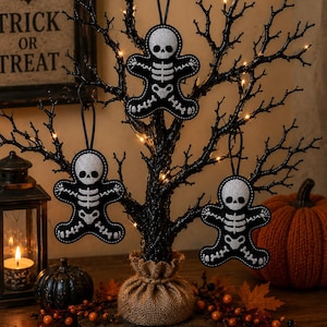 May include: Three black gingerbread-shaped ornaments featuring white embroidered skeleton designs. Each ornament has a circular head, torso, and limbs with stitched bone details and a black hanging loop attached to the top of the head.