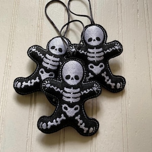 May include: Three black felt gingerbread-style ornaments featuring white embroidered skeleton designs. Each ornament has a rounded head, outstretched arms, and legs, with a black cord loop attached to the top for hanging purposes.