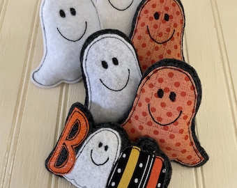 May include: A collection of six plush ghost ornaments made from felt and fabric. Three ghosts are white felt, two are orange with polka dots, and one features the letter B, a white ghost, and a striped pumpkin pattern in black, orange, and yellow.