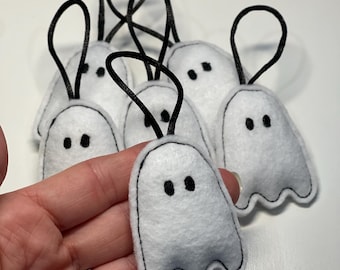Könnte beinhalten: Weiße Filz-Geister-Ornamente mit schwarzen Nahtkonturen und schwarzen Augen. Jeder Geist hat eine schwarze Schlaufe zum Aufhängen. Diese Geister sind eine lustige Dekoration für Halloween.