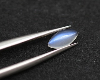 Könnte beinhalten: Ein polierter, ovaler Edelstein mit blau-weißem Farbverlauf, gehalten von einer silbernen Pinzette. Der Edelstein ist glatt und reflektierend, vor einem dunklen, strukturierten Hintergrund.