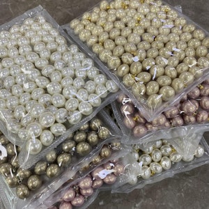 May include: A close-up of three bags of white, cream, and pink pearl beads. The beads are arranged in a variety of sizes and shapes.