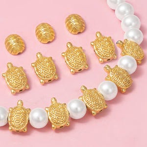 May include: Gold-colored turtle-shaped beads and oval beads with textured shell patterns. The beads are interspersed with white pearl beads, creating a decorative jewelry element. The background is a soft pink.