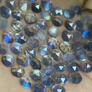 May include: A collection of iridescent blue-grey gemstone beads, each with a faceted, rounded shape. The beads are arranged in a loose pile on a hand.