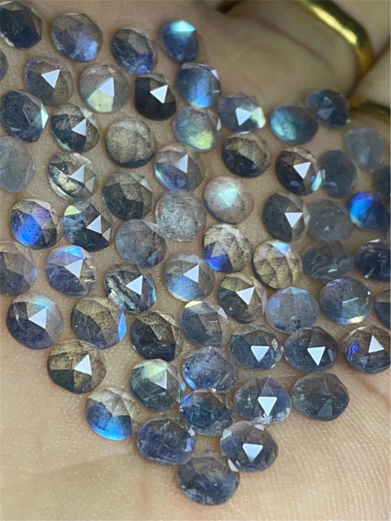 May include: A collection of iridescent blue-grey gemstone beads, each with a faceted, rounded shape. The beads are arranged in a loose pile on a hand.