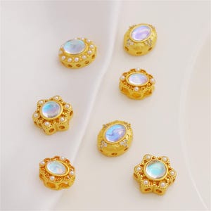 May include: Six gold-toned metal beads featuring central iridescent oval cabochons. The beads are accented with small white pearls and clear crystals, with some designs shaped like flowers and others as oval frames with side perforations.
