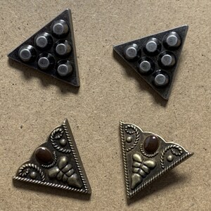 May include: Four metal triangle corner protectors. Two are silver with a hammered finish and six round studs. Two are gold with a decorative design and a brown stone in the center.