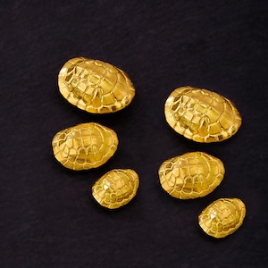 May include: Six gold-colored, oval-shaped decorative objects with a textured, segmented surface, resembling turtle shells. The objects are arranged on a dark background, creating a contrast that highlights their metallic sheen.