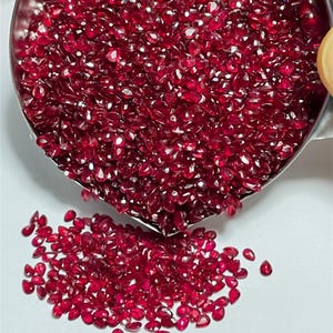 Könnte beinhalten: Nahaufnahme einer großen Menge loser, facettierter, birnenförmiger, roter Edelsteine. Die Edelsteine befinden sich in einer Metallschale und fallen auf eine weiße Oberfläche.