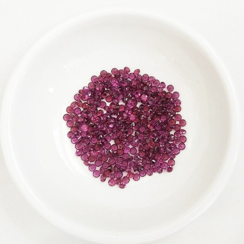 May include: A pile of small, round, pink gemstones in a white bowl. The gemstones are all about the same size and are arranged in a random pattern.