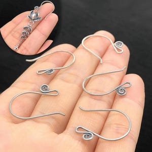 May include: Silver-toned earring hooks with a looped design. The image shows several individual hooks, with a close-up of a finished earring with a floral and leaf design. The hooks are made of a shiny, metallic material.