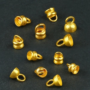 May include: A selection of small, gold-coloured metal jewellery findings. Each piece has a bell-like shape with a small loop attached. The findings are scattered on a dark grey surface.