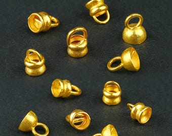 May include: A collection of small, gold-colored metal jewelry findings. Each piece has a bell-like shape with a small loop attached. The findings are scattered on a dark gray surface.