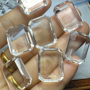 Könnte beinhalten: Eine Sammlung von acht klaren, facettierten, rechteckigen Quarzkristallen. Die Kristalle sind auf einer Hand angeordnet, wobei sich einige überlappen.