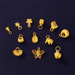 Op de afbeelding: Een verzameling kleine, goudkleurige bedels in verschillende vormen, waaronder een microfoon, bloem, kam, olifant en vlinder. Elke bedel heeft een lus om aan een ketting of armband te bevestigen. De bedels worden gepresenteerd tegen een donkerblauwe achtergrond.