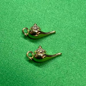 以下が含まれることがあります： 宝石をちりばめた上部と湾曲した注ぎ口が付いた、2つの金色のジーニーランプ。ランプは緑色の背景にあります。ランプには小さなハンドルと詳細なデザインがあります。