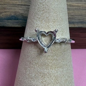 May include: A silver ring with a heart-shaped setting. The ring features small, clear stones along the band and prongs for securing a gemstone. The ring is displayed on a neutral-toned cone.