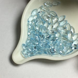 May include: A white scoop filled with a large quantity of light blue marquise shaped gemstones.