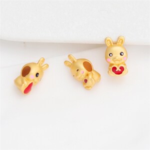 Peut inclure: Trois petites breloques en forme de lapin de couleur dorée, chacune avec une finition mate, des accents de joues roses et un détail en forme de cœur rouge vif tenu au centre. Les figurines de lapin sont représentées dans diverses positions.