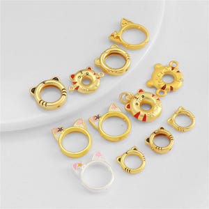 Può includere: Una collezione di anelli e ciondoli in metallo color oro a forma di teste di gatto. Alcuni pezzi presentano accenti in smalto rosa, viola e rosso, mentre altri sono in oro semplice. I design includono anelli circolari con orecchie a punta.