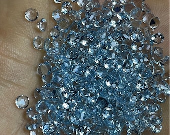 May include: A large pile of light blue aquamarine gemstones, also known as aquamarine crystals, on a hand. The gemstones are round and faceted.